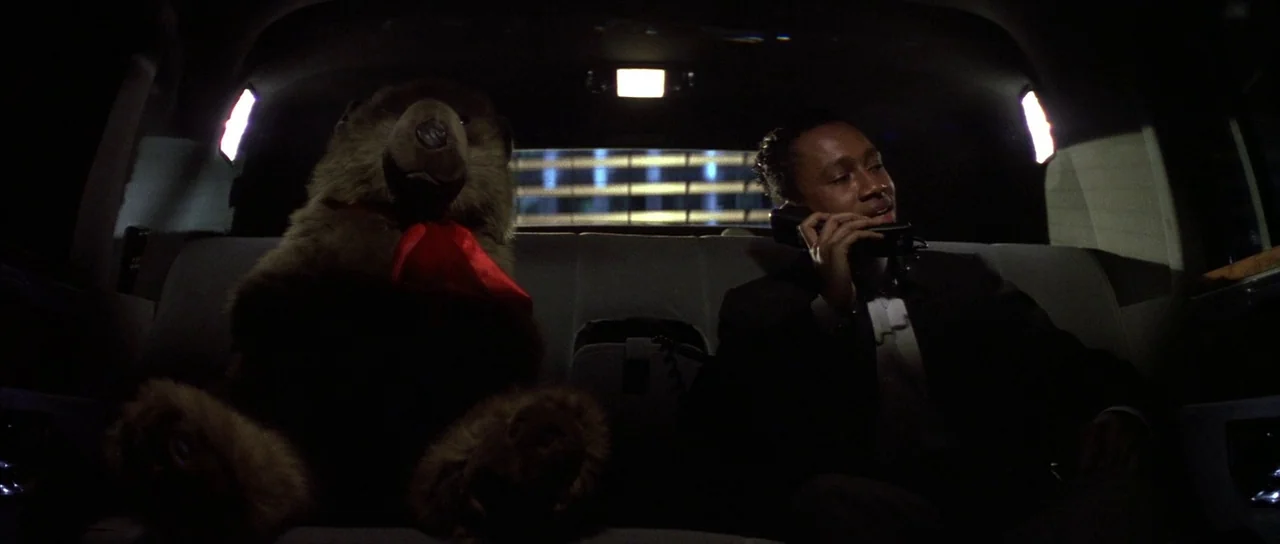Die Hard (1988) movie still: medium — The interior of a darkened luxury vehicle with gray leather seats. A man in a suit sits beside a large stuffed…