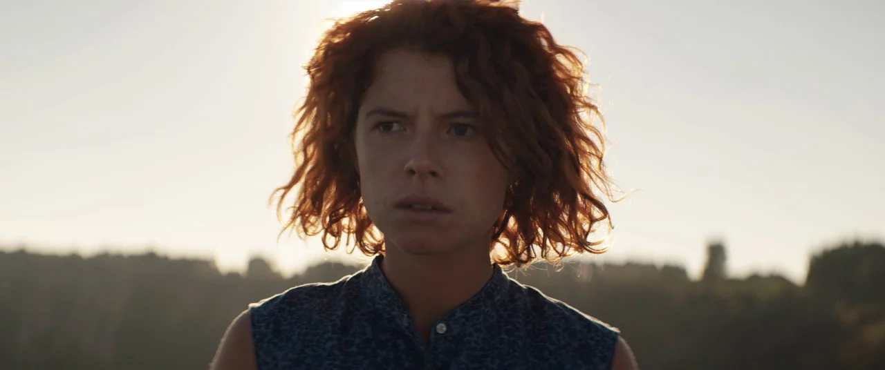 Beast (2017) movie still: medium — A woman stands in a rural landscape during the golden hour sunset. She faces the camera while the low sun…