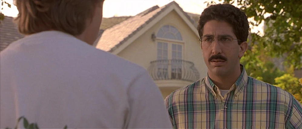 Apt Pupil (1998) movie still: medium — A residential setting shows a light-colored house with a small balcony in the distance. A man wearing a plaid…