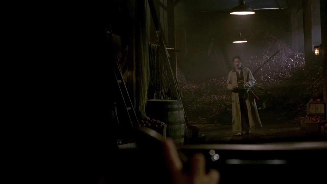 In Dreams (1999) movie still: wide — A dark, cavernous interior space contains large piles of produce and wooden crates. A man in a trench coat…