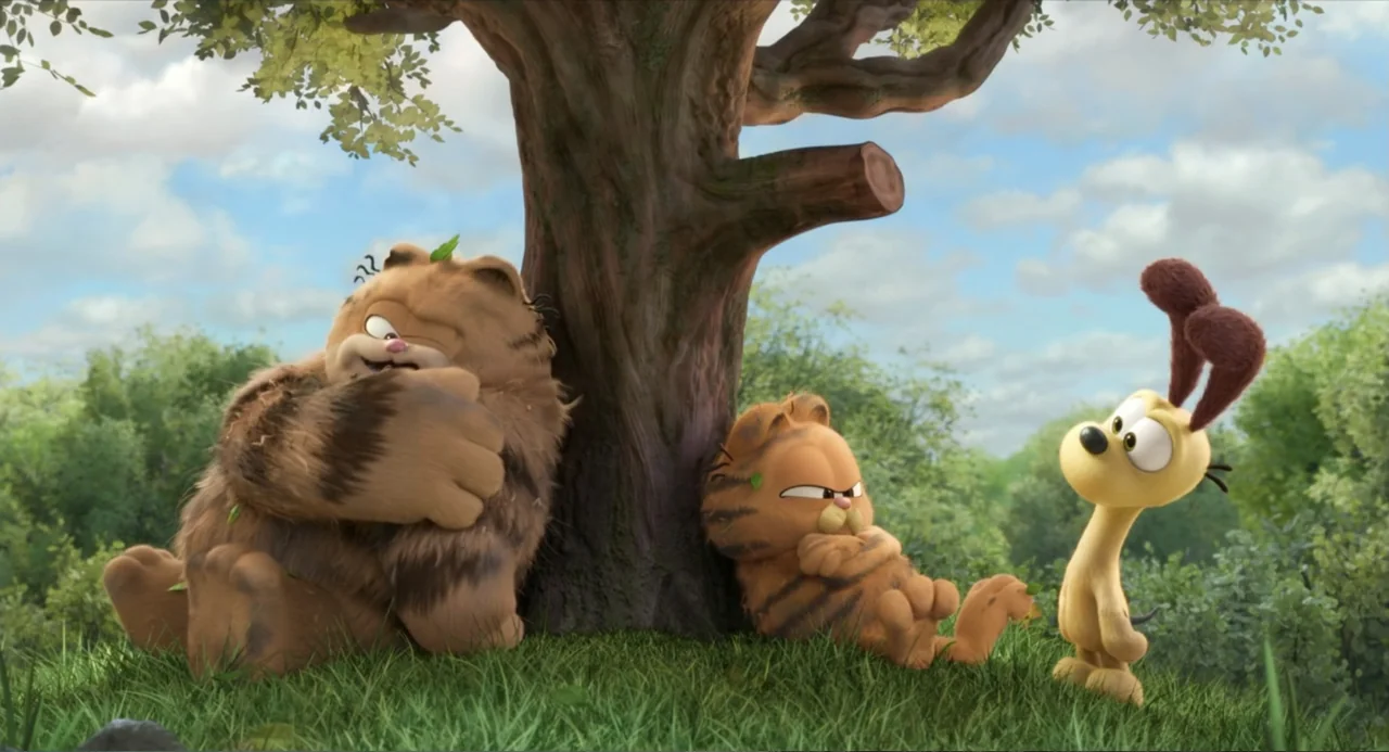 The Garfield Movie (2024) movie still: medium — A large, gnarled tree stands in a bright, grassy field under a cloudy sky. Two orange cats recline…