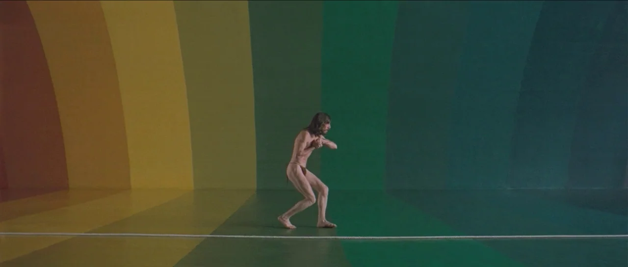 The Holy Mountain (1973) movie still: wide — A man stands on a rope in front of a wall with vertical rainbow stripes. The floor matches the wall…