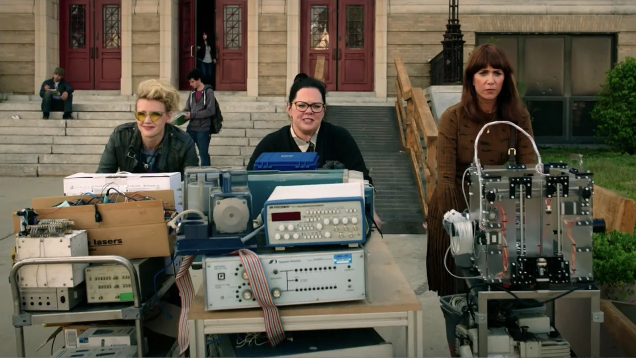 Ghostbusters (2016) movie still: medium — Three women stand behind metal tables containing various electronic test equipment and circuit components.…