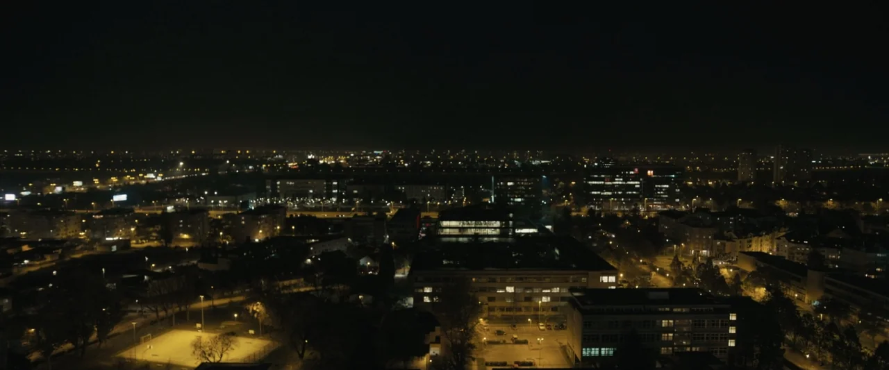 Canary Black (2024) movie still: establishing — A wide aerial view of a sprawling urban landscape at night with buildings. Multiple illuminated…