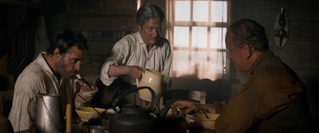 The Sisters Brothers (2018) movie still: medium — A rustic wooden room contains a dining table with ceramic bowls and a metal kettle. Three people…