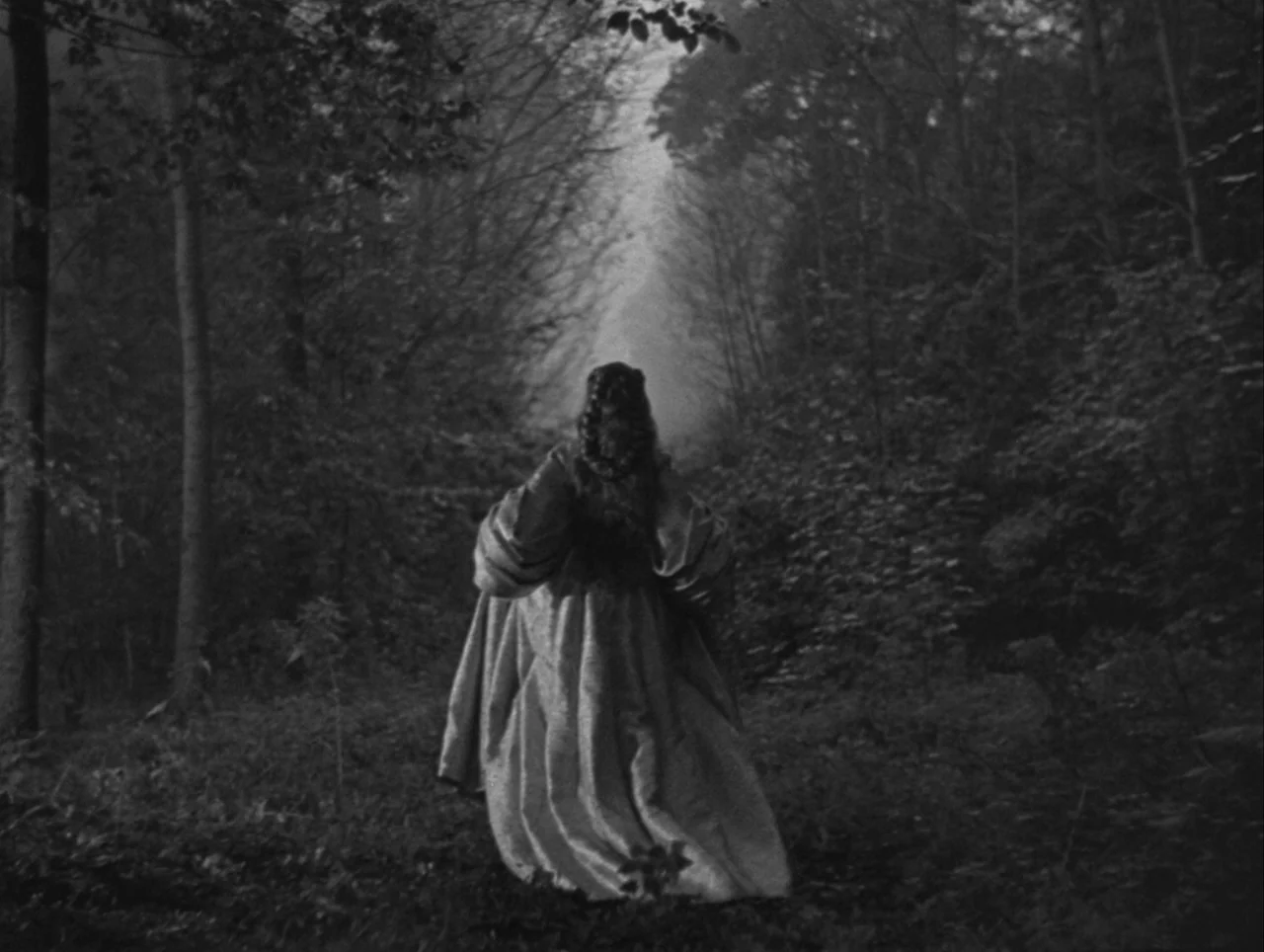 La Belle et la Bête (1946) movie still: A solitary figure in a flowing, historical gown walks away from the camera, down a narrow, overgrown forest…
