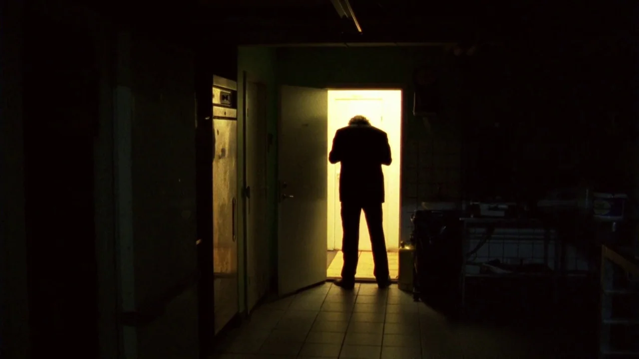 Pusher III (2004) movie still: fullbody — A dimly lit utility room with tiled floors and dark, cluttered surfaces. A lone man in a dark suit stands…