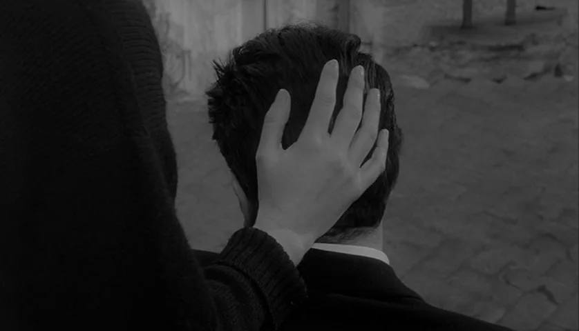 L’Avventura (1960) movie still: closeup — An outdoor paved area with a stone wall visible in the background. A woman reaches out to touch the hair…
