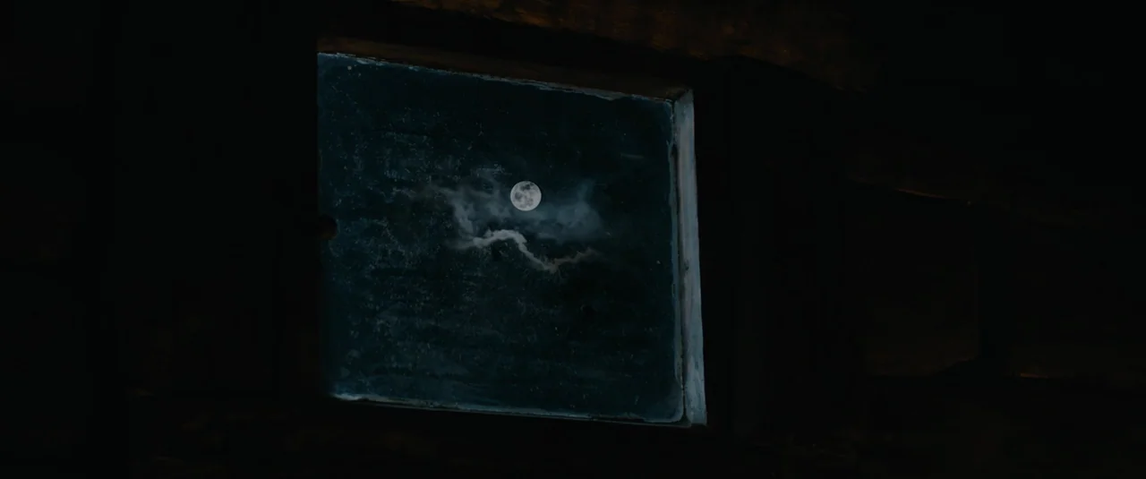 Hagazussa (2017) movie still: wide — A small square window is set into a dark wooden wall of a building. The moon shines brightly through the window…