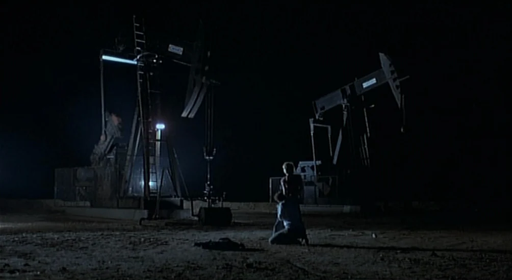 Near Dark (1987) movie still: wide — An expanse of dark, barren dirt ground is punctuated by two mechanical oil pumps. Two men stand in the middle…