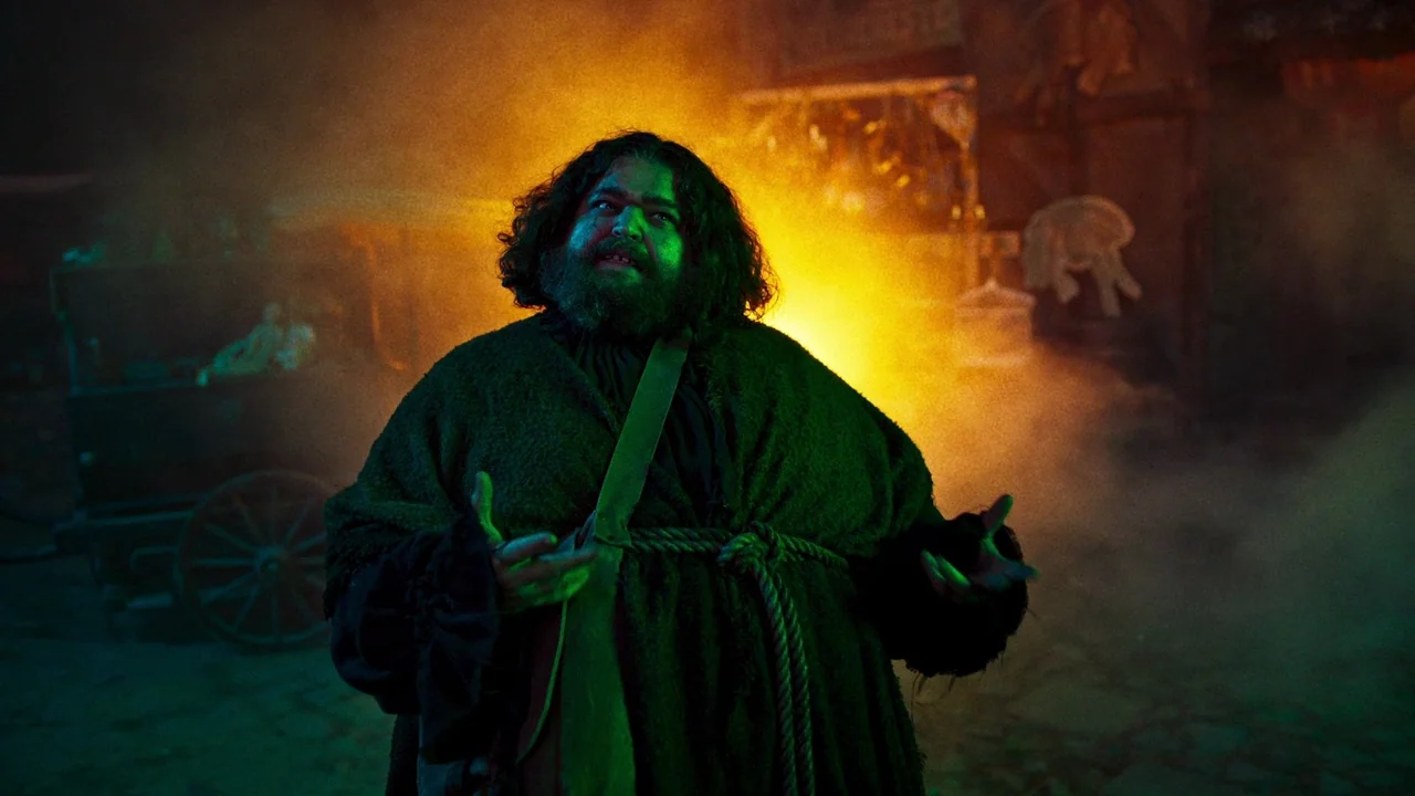 The Munsters (2022) movie still: medium — A bustling market square at night is filled with thick fog and yellow smoke. A bearded man in a heavy dark…