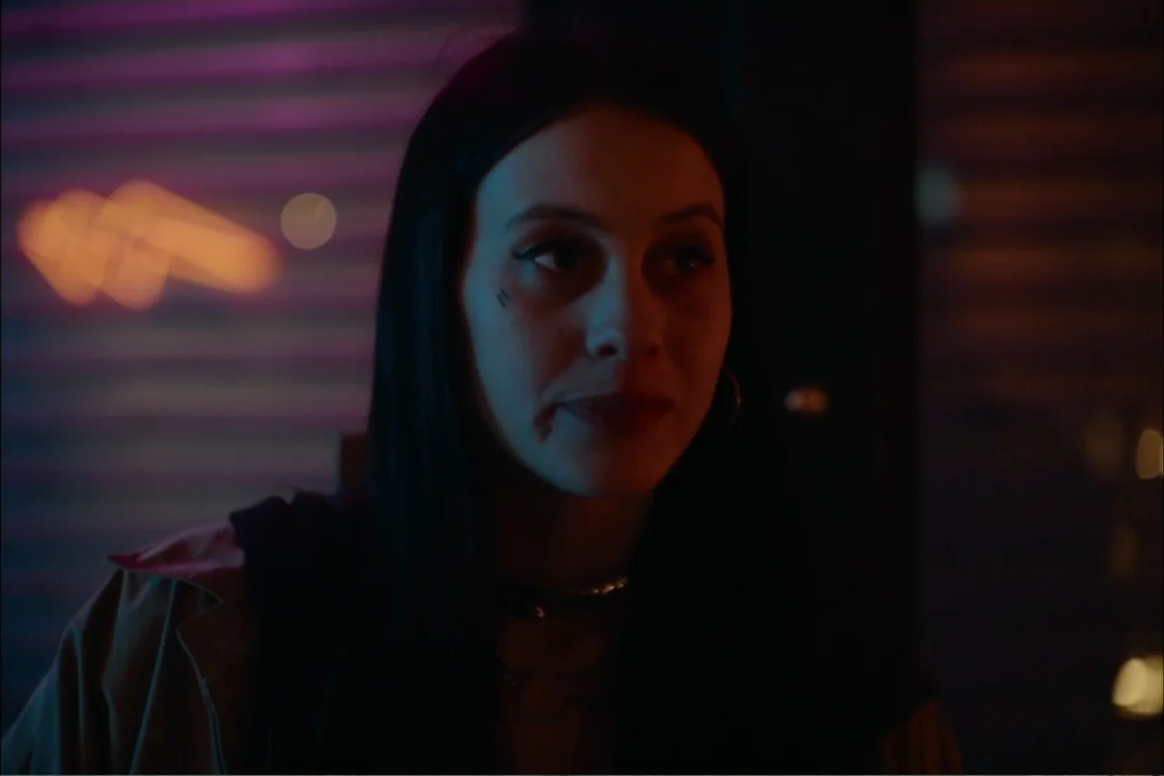 Cross the Line (2020) movie still: closeup — A dark indoor space with blurred neon lights visible in the background reflection. A young woman with…