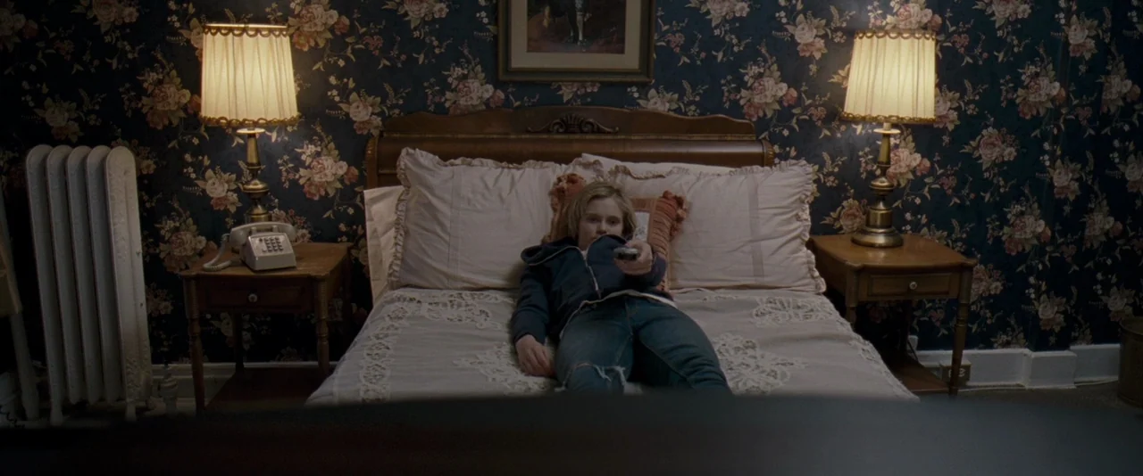 The Innkeepers (2011) movie still: medium — A bedroom features floral wallpaper and a wooden bed frame with beige linens. A teenage girl lies…