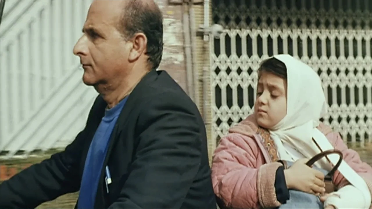 The Mirror (2014) movie still: medium — A man and a young girl ride a motorcycle in front of metal shutters. The man operates the bike while the…