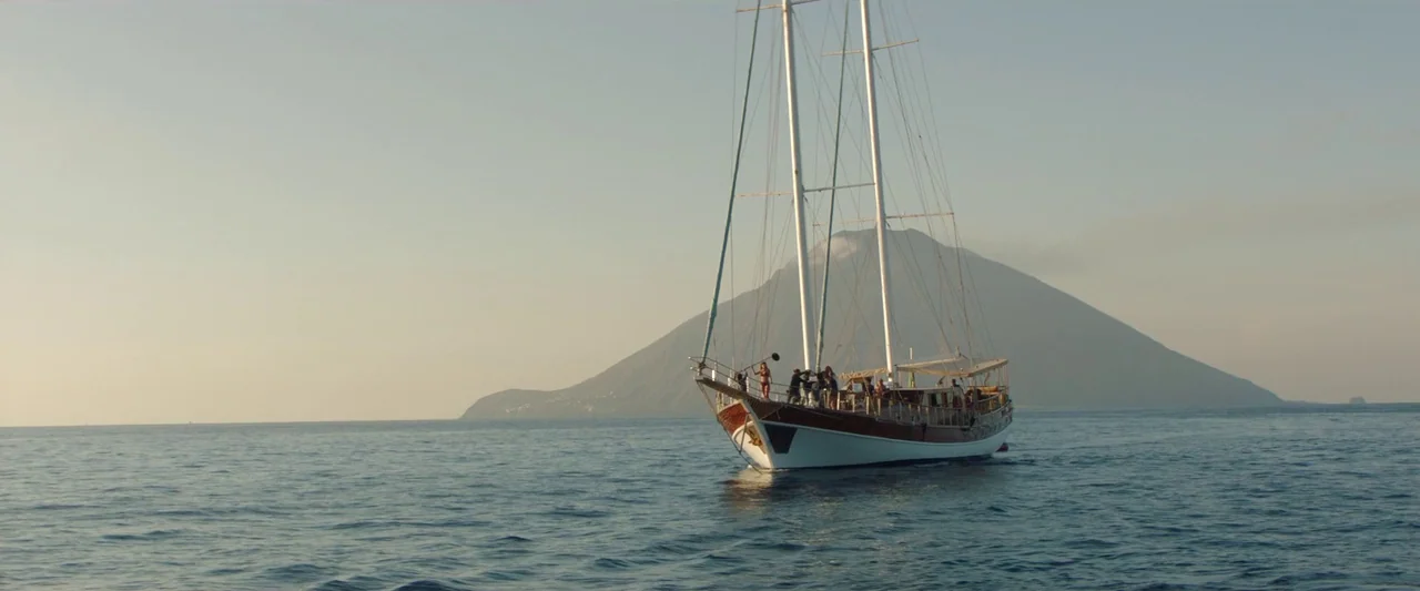 Sibyl (2019) movie still: wide — A wooden gulet sailboat travels across calm water toward a large volcanic island. Several passengers stand on the…