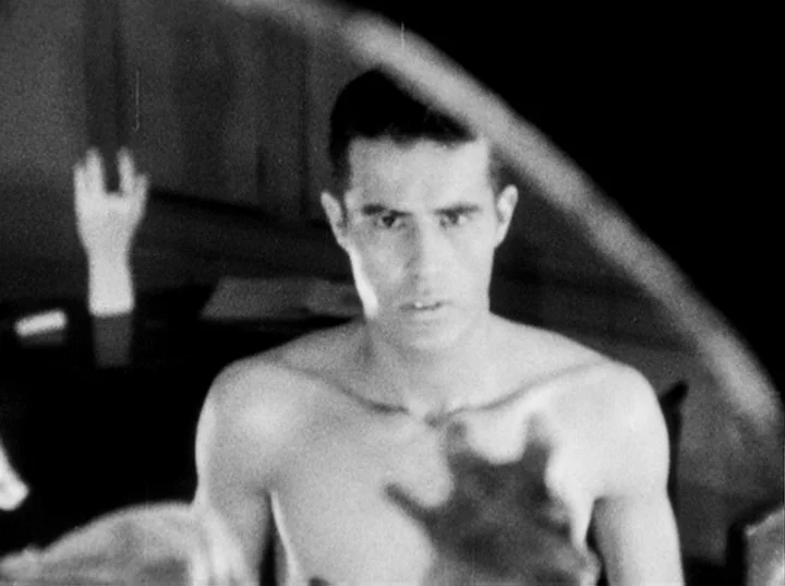 Fireworks (1947) movie still: closeup — A dimly lit space containing a reflective glass surface and a mannequin hand. The shirtless man stares…