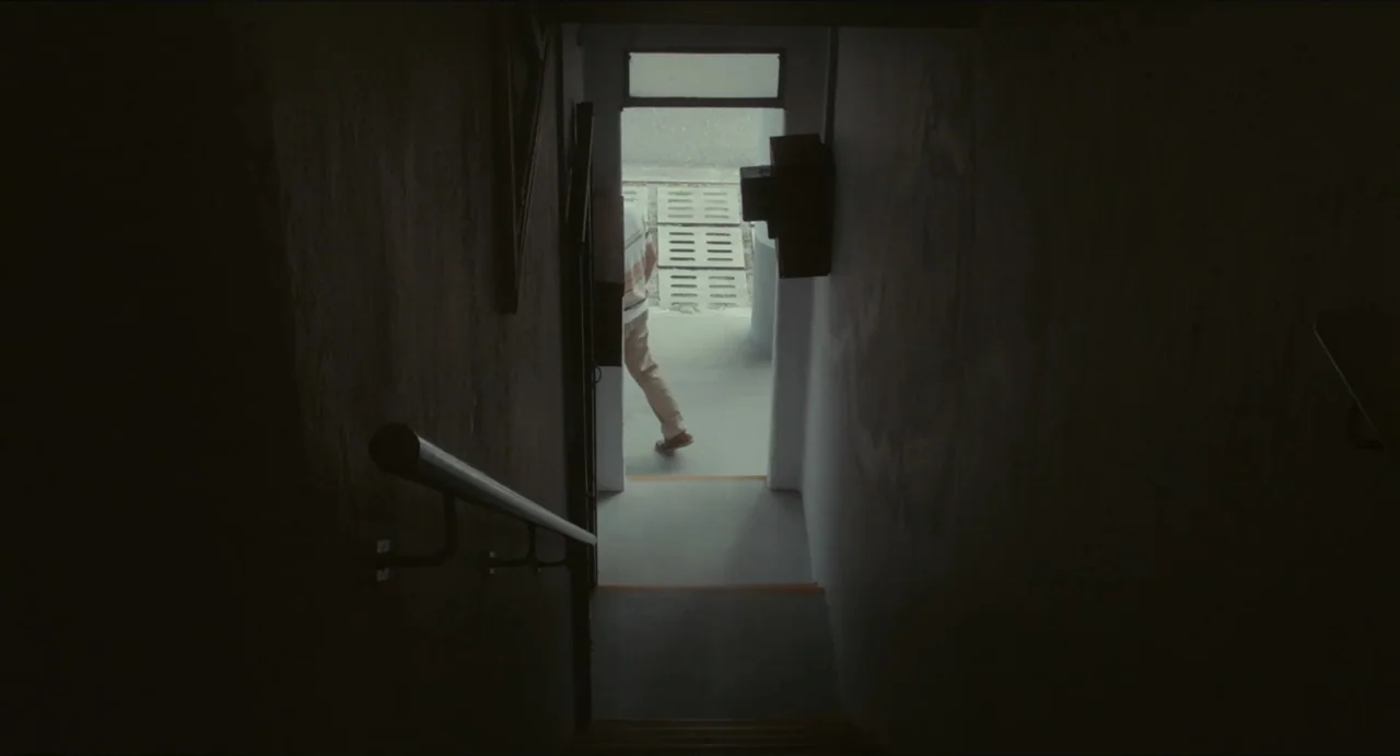 Ramen Shop (2018) movie still: medium — A dark, enclosed stairwell leads toward a brightly lit exterior doorway at the base. A man in a striped…