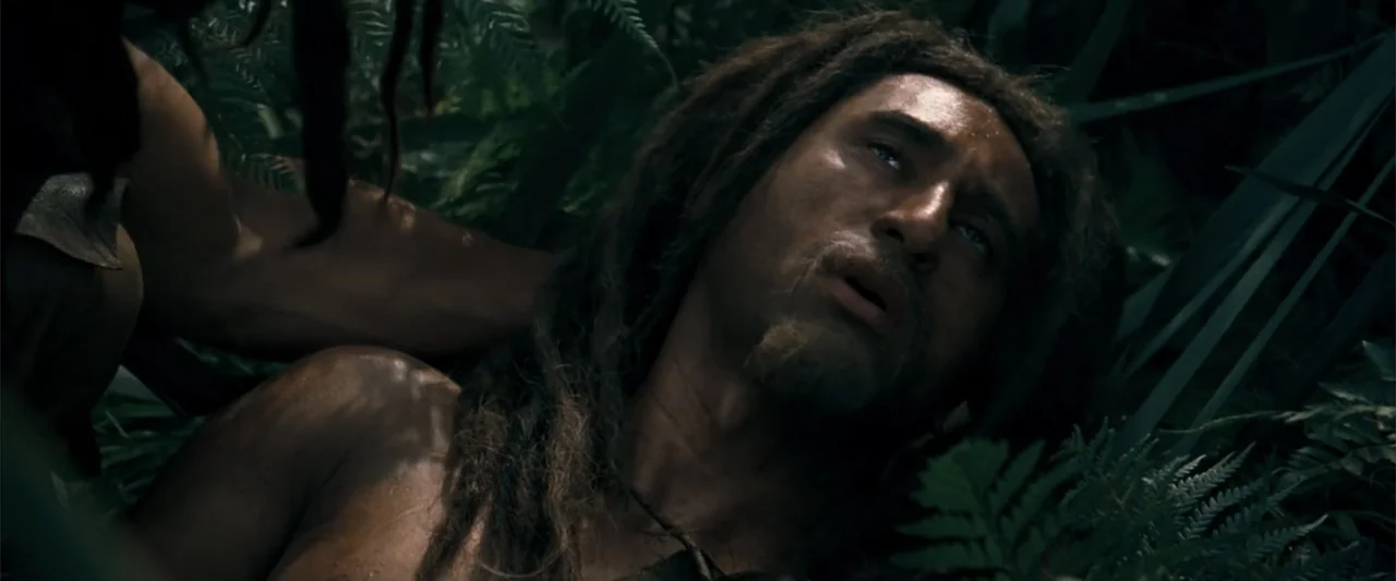 10,000 BC (2008) movie still: closeup — A dense jungle floor with thick ferns and shadowed foliage. A man with dreadlocks lies on the ground, gazing…