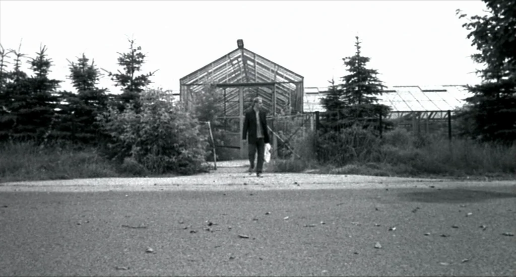 Run Lola Run (1998) movie still: wide — A man walks away from a glass-walled structure surrounded by evergreen trees and brush. An asphalt path…