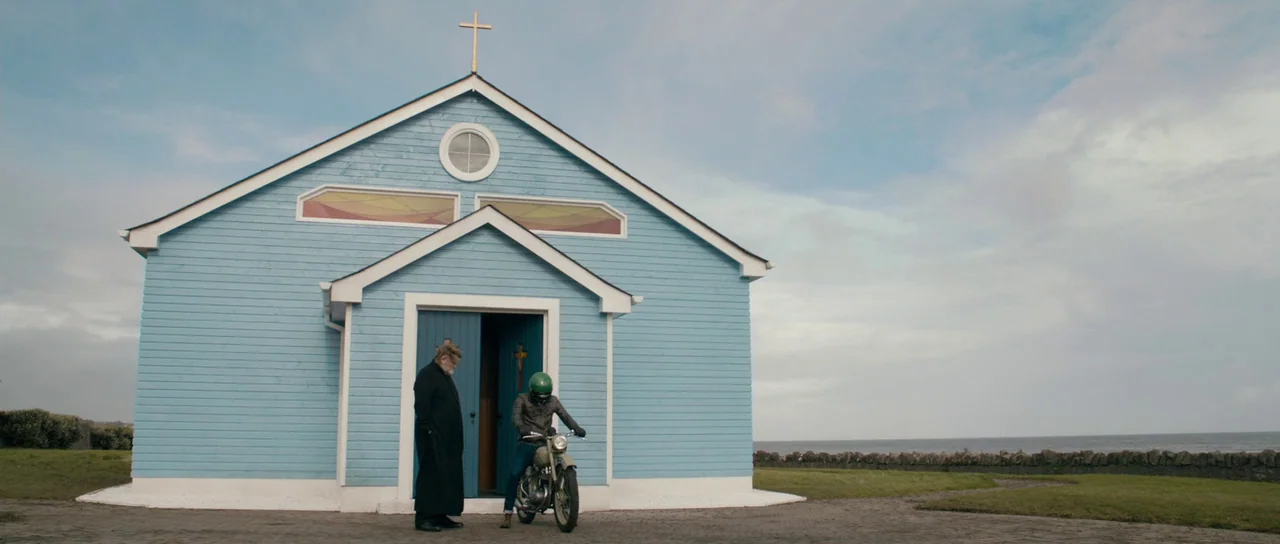 Calvary (2014) movie still: wide — A bright blue wooden building with a cross sits on a grassy field. A priest talks to a motorcyclist parked in…
