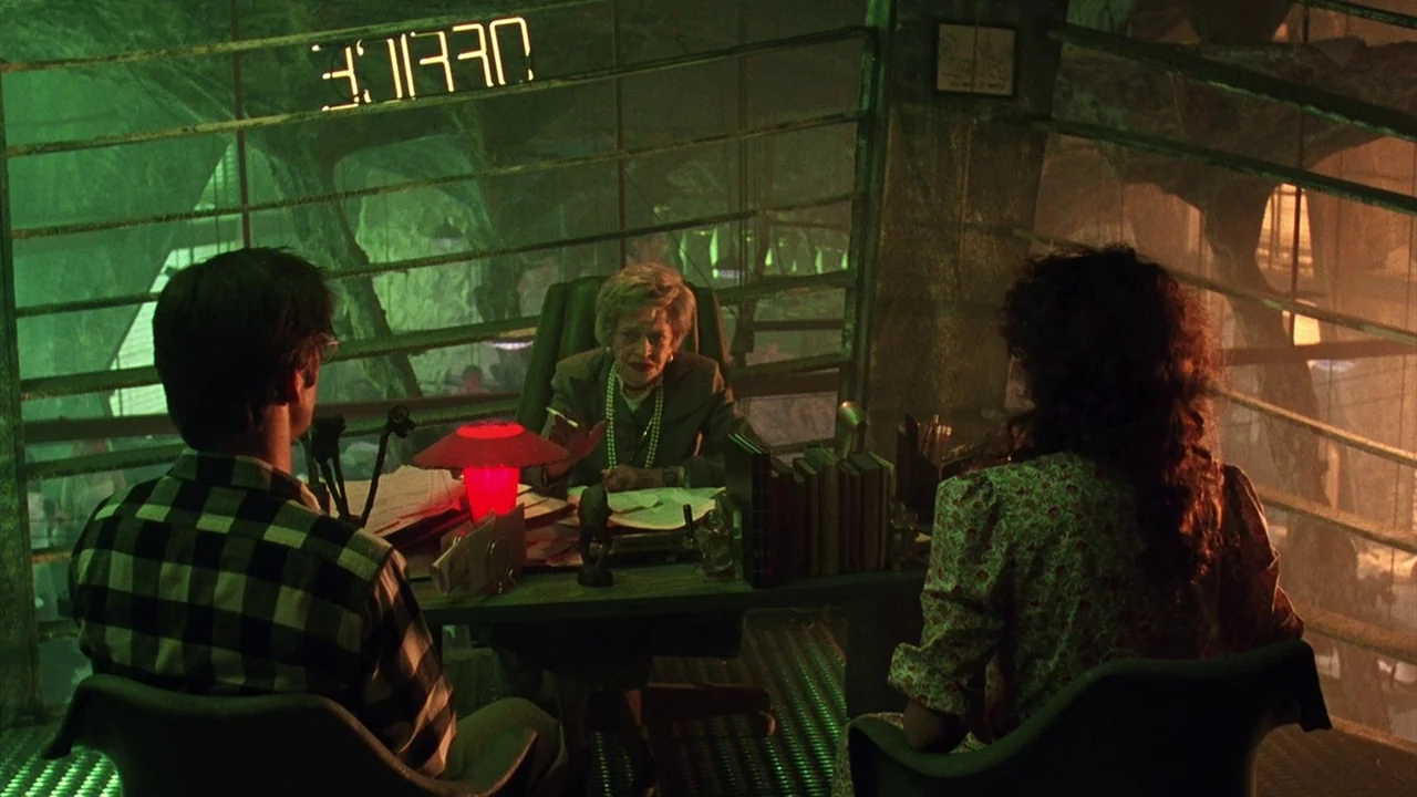 Beetlejuice (1988) movie still: medium — An industrial-style office room features metal grates, green lighting, and a backwards neon sign. Two…
