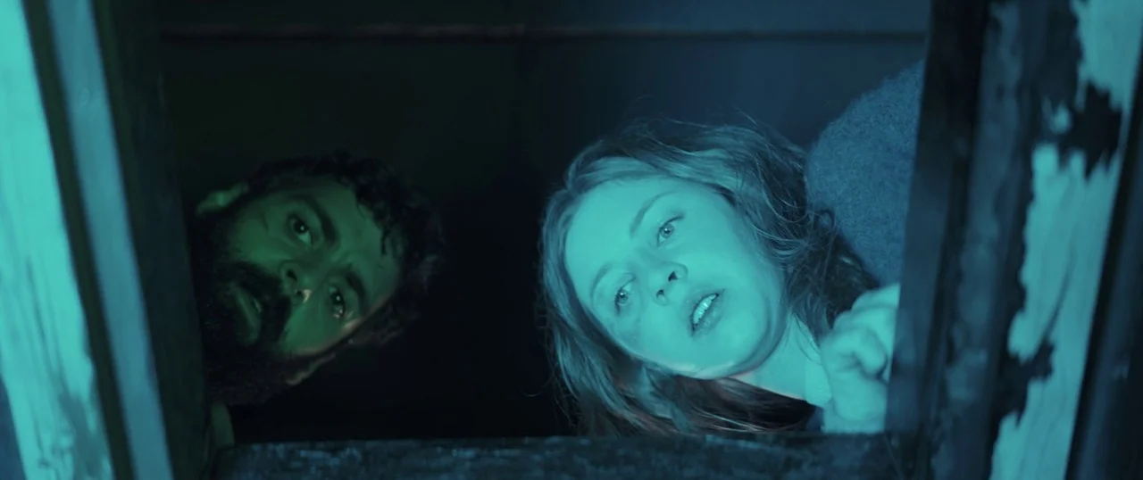 Sea Fever (2019) movie still: medium — A dark, cramped space frames the faces of a man and woman looking down. The lighting highlights their…