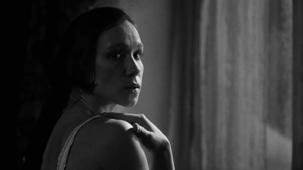 The Role (2013) movie still: closeup — A dark bedroom interior with a draped window partially visible in the background. A woman in a tank top turns…