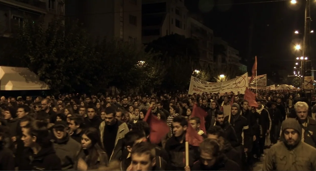 Az X- Kryptonit projekt (2020) movie still: wide — A dense crowd of people marches down a night-time city street lined with tall buildings. They…