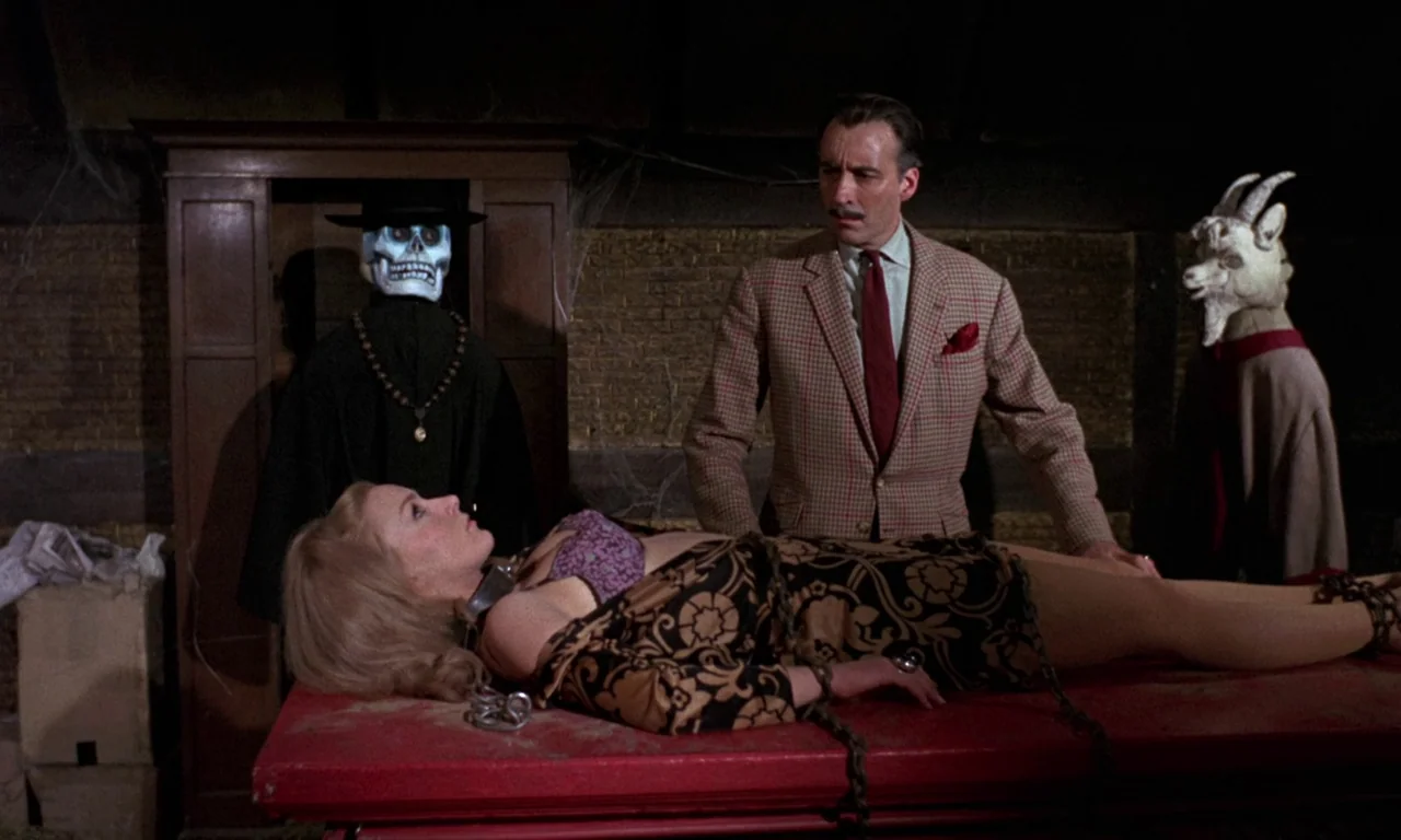 Curse of the Crimson Altar (1968) movie still: medium — A dimly lit wooden space containing a skull-masked figure and a goat-headed figure. A woman…