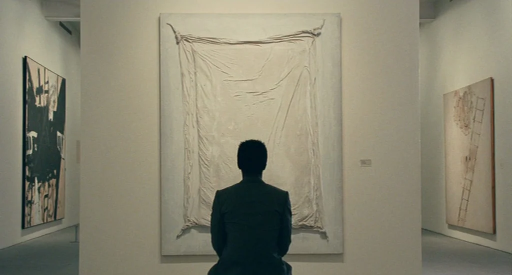 The Limits of Control (2009) movie still: medium — A minimalist art gallery with white walls displaying abstract paintings. A man sits on a bench…