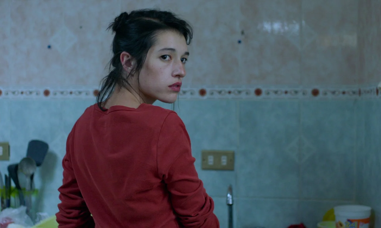 The Untamed (2016) movie still: medium — A tiled room with light blue wall tiles and a horizontal decorative border. A young woman in a red shirt…