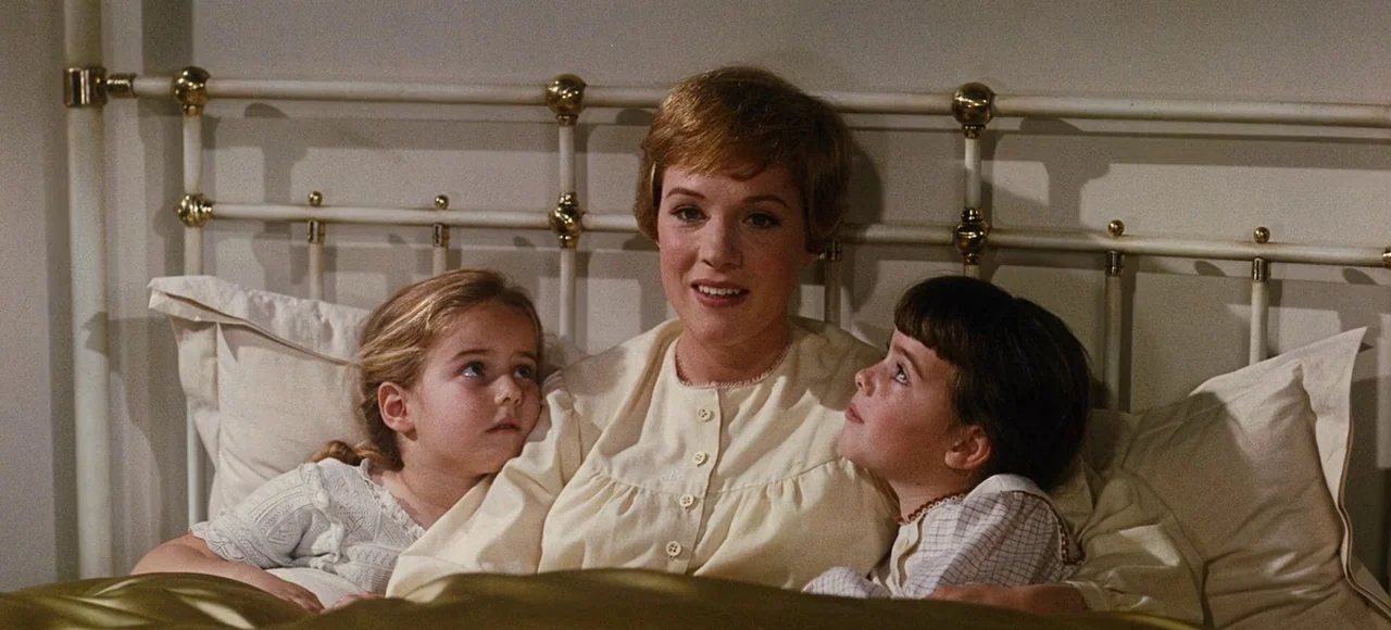 The Sound of Music (1965) movie still: medium — A woman and two young children sit together in an iron-framed bed. They are resting against large…