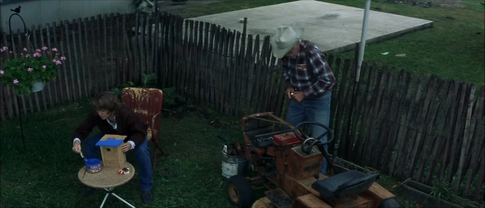 The Straight Story (1999) movie still: wide — A grassy backyard features a wooden fence, a small ride-on mower, and potted flowers. A woman sits on…