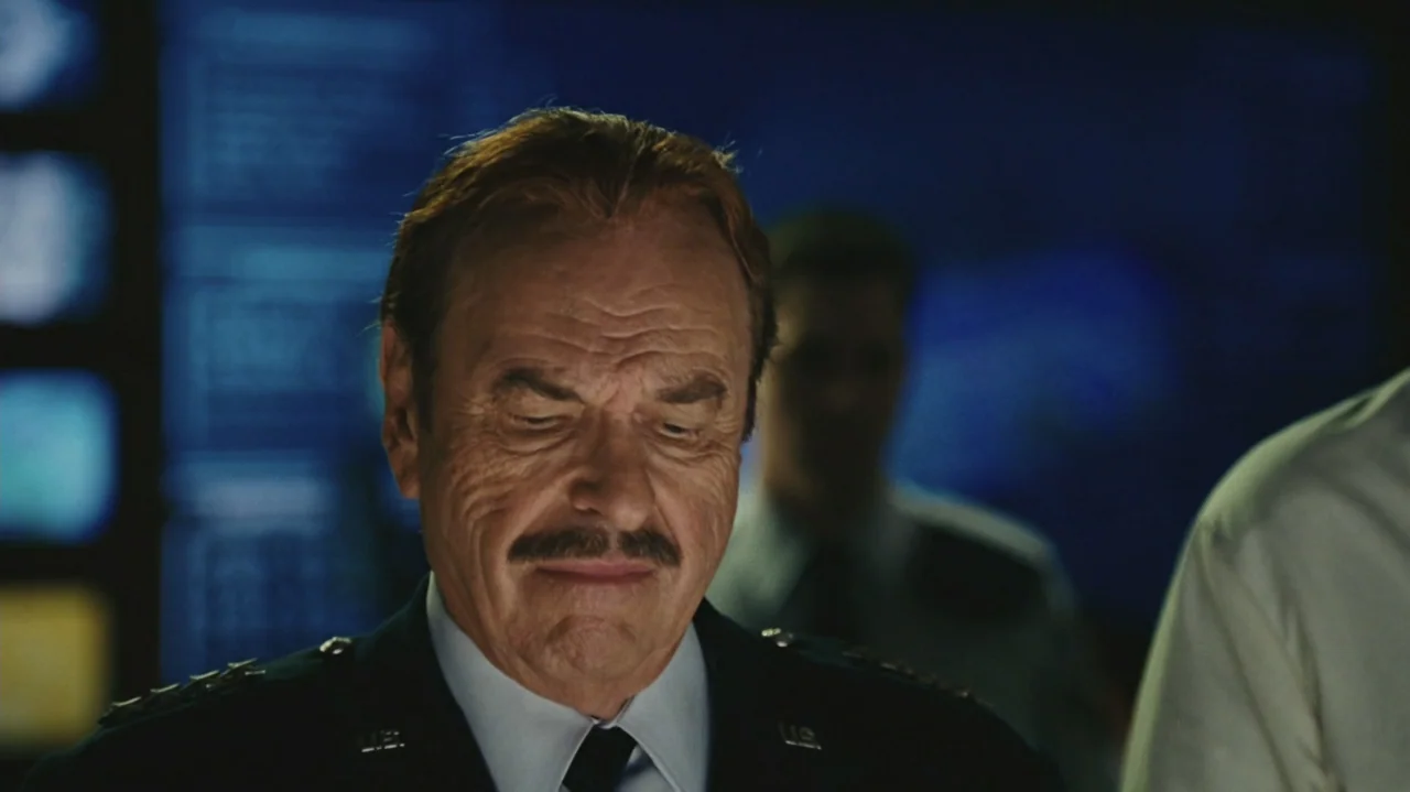 Zoom (2006) movie still: closeup — A dimly lit command center with large dark digital displays in the background. An older military officer stands…