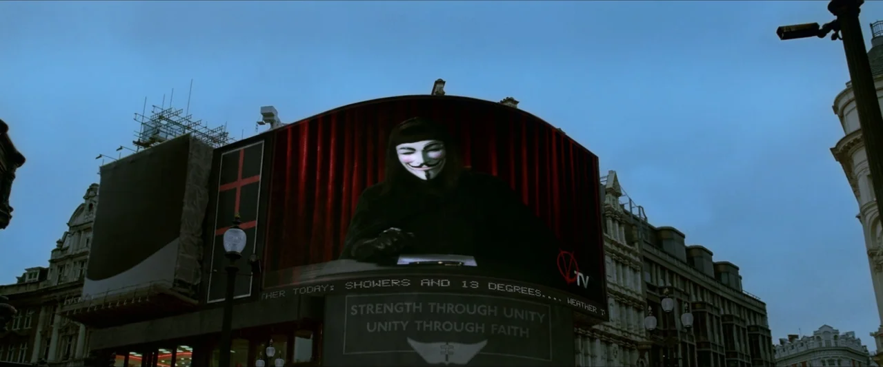 V for Vendetta (2006) movie still: wide — A large public screen displays a masked figure to an empty city street at dusk. The screen text reads…