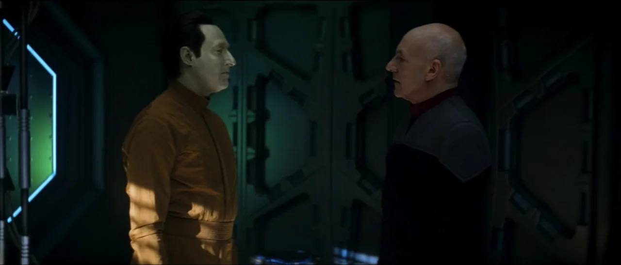 Star Trek: Nemesis (2002) movie still: medium — A dark, metallic futuristic interior features paneled walls and inset lighting fixtures. Two men in…