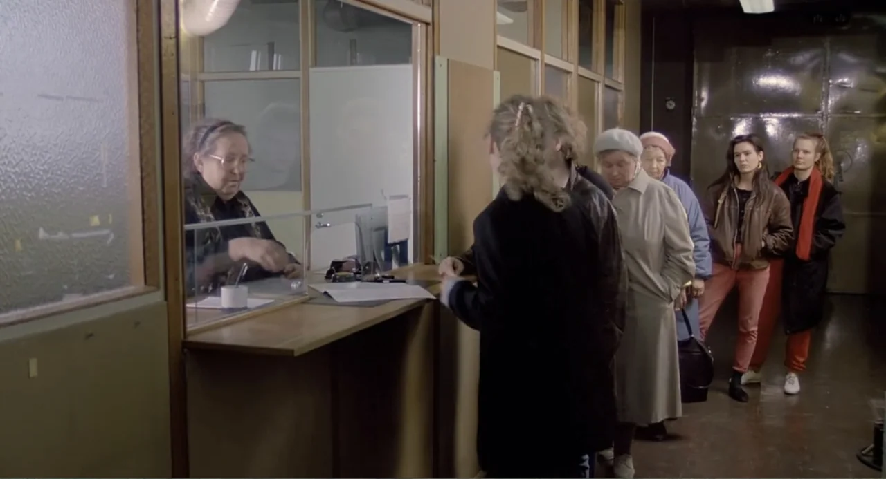 The Match Factory Girl (1990) movie still: medium — An interior service window with light wood panels and glass partitions separates a clerk. A line…
