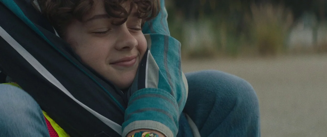 Honey Boy (2019) movie still: closeup — A person with curly hair rests their head against another individual in an outdoor space. The background is…