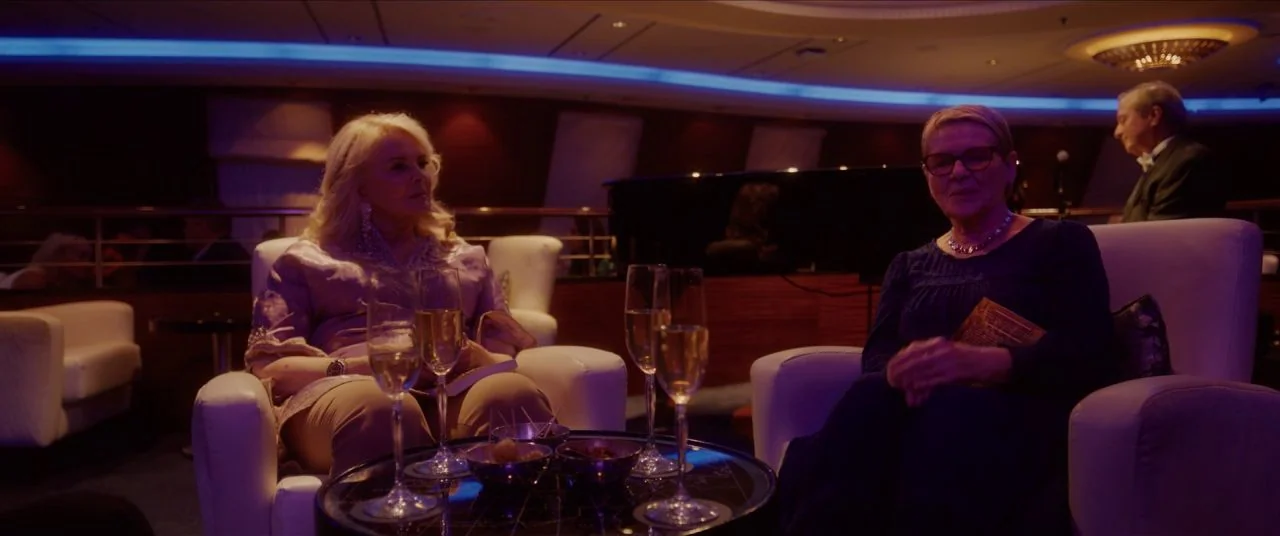 Let Them All Talk movie still: medium — An dimly lit lounge with white chairs and blue LED accent lighting. Two women sit in white armchairs and…