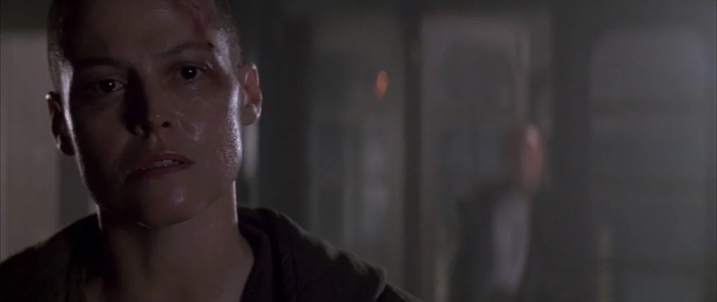 Alien 3 (1992) movie still: closeup — A dimly lit, metallic industrial setting with shadowed structures visible in the background. A woman with a…