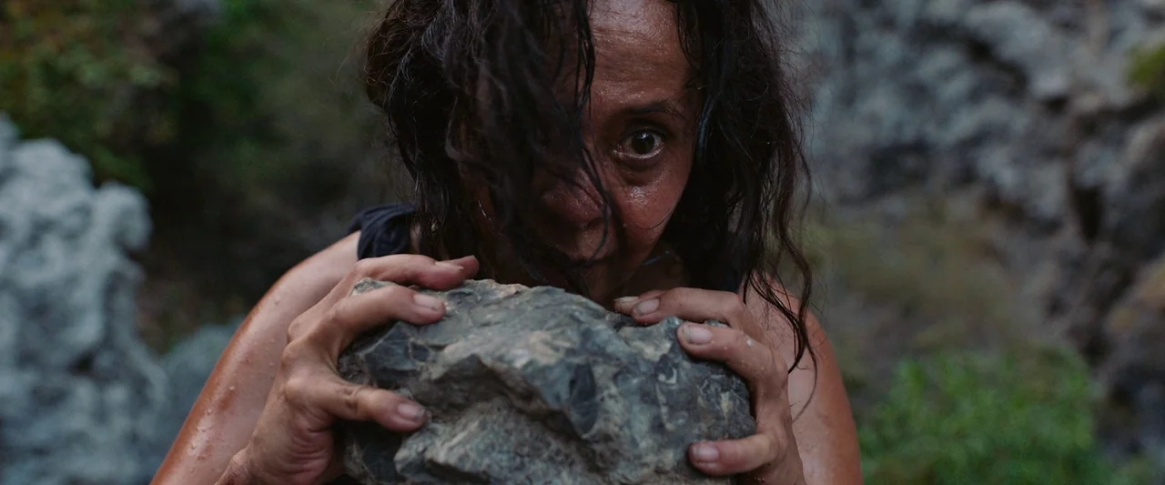 Triangle of Sadness (2022) movie still: closeup — A rugged exterior environment features sharp grey stone and patches of low-lying greenery. A woman…