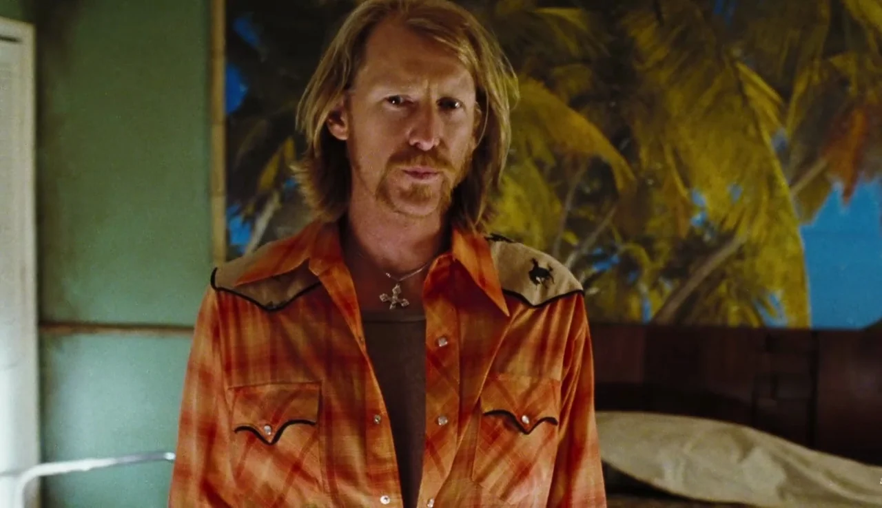 The Devil’s Rejects (2005) movie still: medium — A man stands inside a room with a wall mural depicting palm trees. He is centered in the frame…