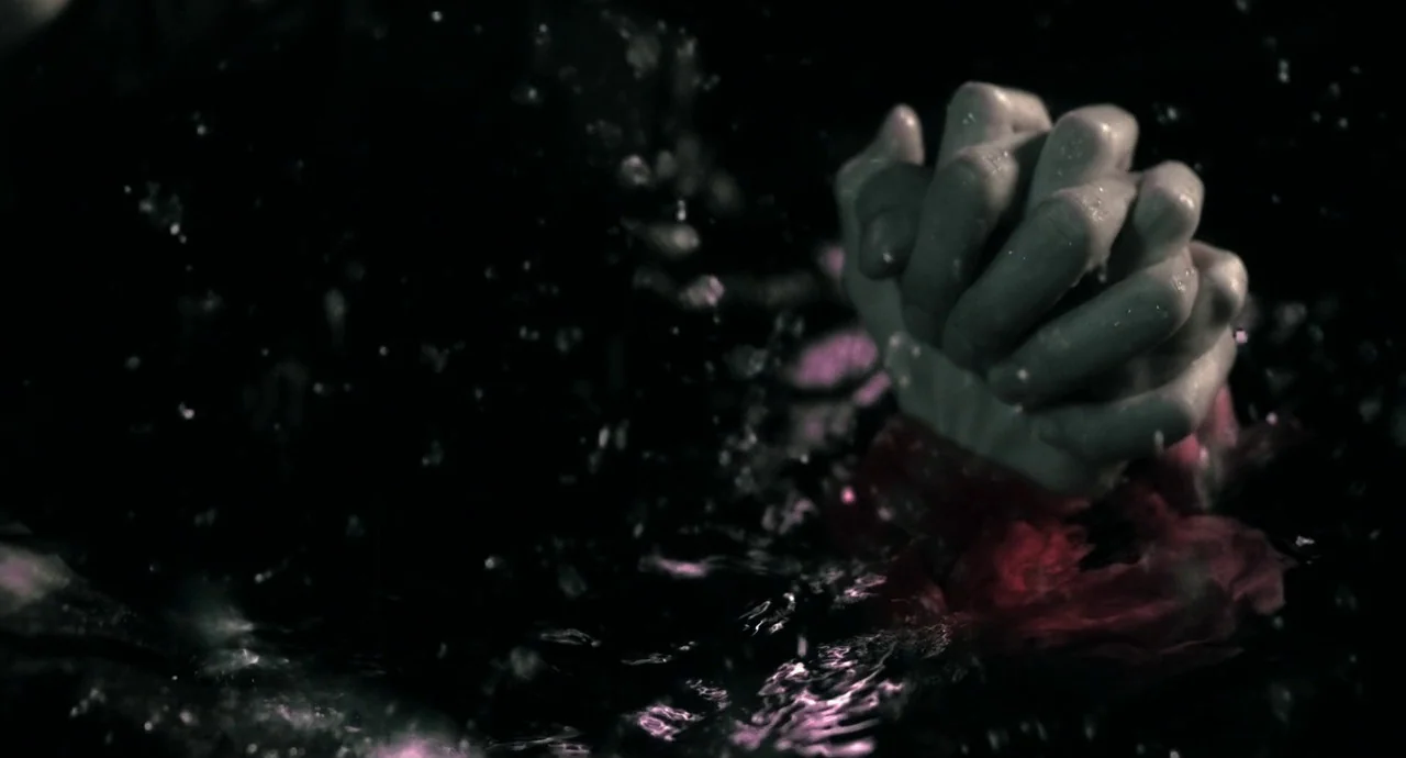 No Longer Human (2019) movie still: closeup — Dark water ripples violently around a pair of pale, clasped hands. Red pigment or blood disperses into…