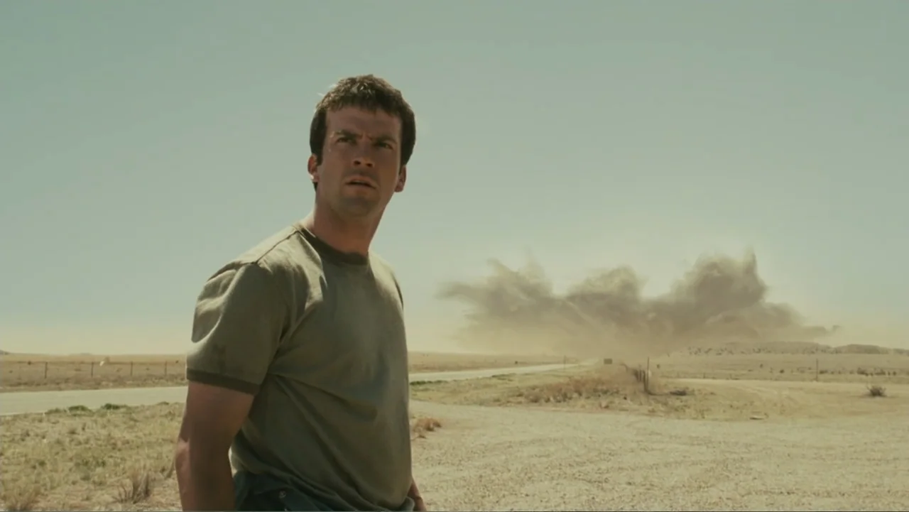 Legion (2010) movie still: medium — A flat desert landscape stretches out under a hazy sky with a distant explosion. A man stands in the foreground…