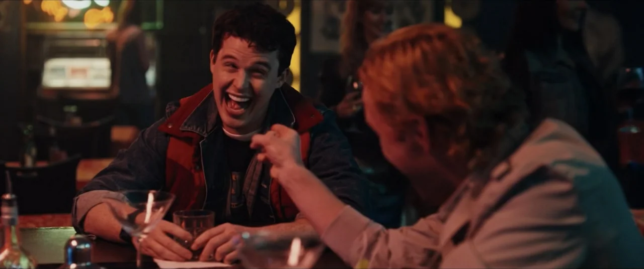 Vicious Fun (2021) movie still: medium — A dimly lit bar interior featuring a wood grain counter and dark booths. A young man sits at the bar…