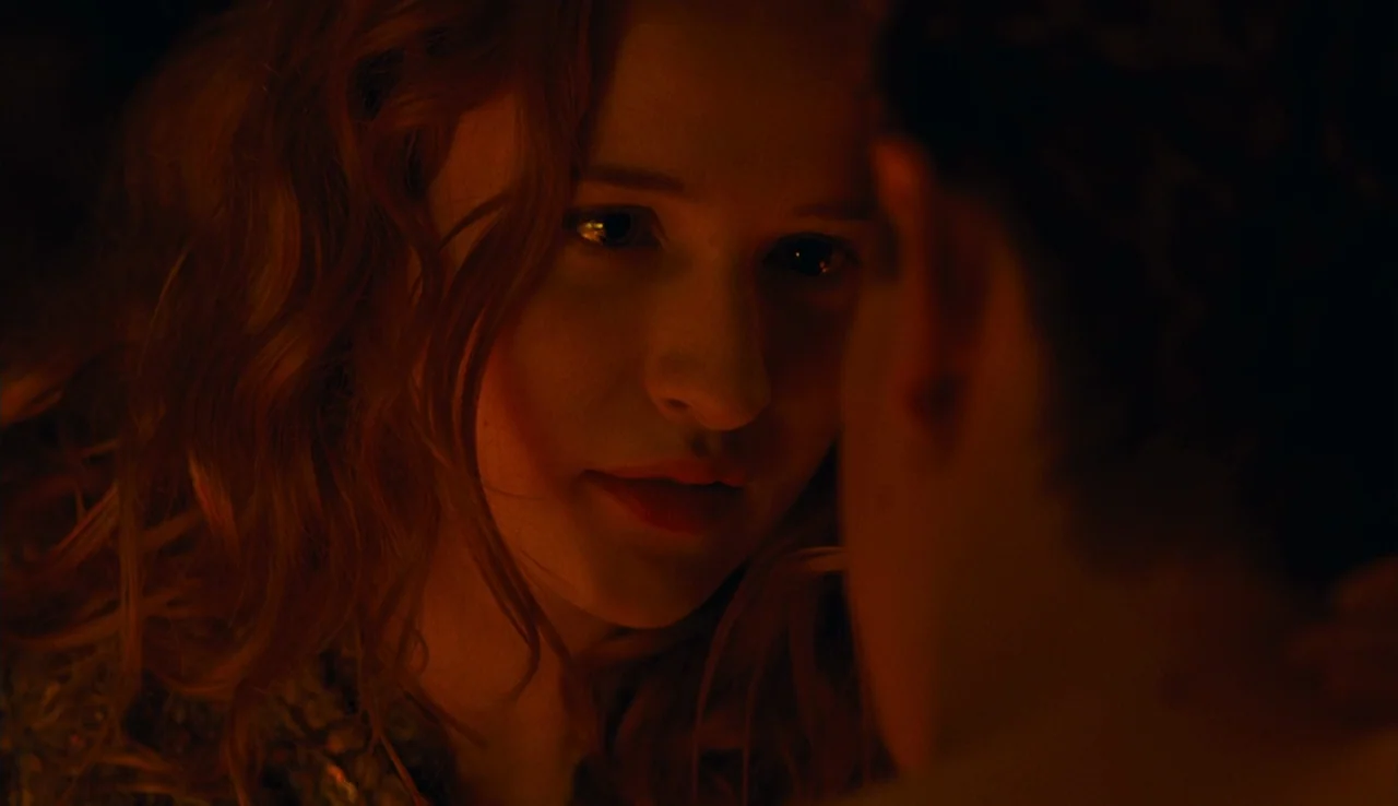 Renoir (2012) movie still: closeup — A dark space lit primarily by an off-screen, warm light source. A woman with long wavy red hair looks directly…