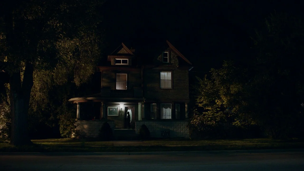 Girl on the Third Floor (2019) movie still: wide — A dark two-story Victorian-style house stands at night behind a stretch of dark lawn. Yellow…