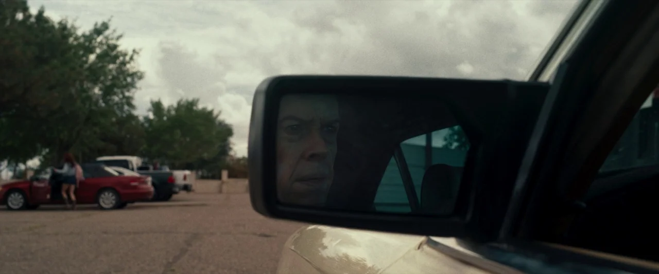 LaRoy, Texas (2023) movie still: closeup — An asphalt parking lot under an overcast sky contains several parked passenger vehicles. The reflection…