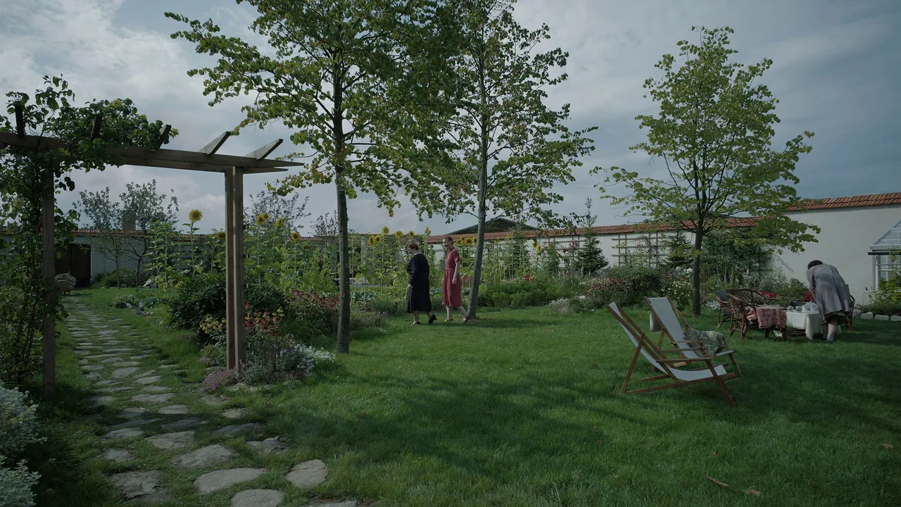 The Zone Of Interest (2023) movie still: wide — A green lawn surrounded by sunflowers and tall trees features a stone walking path. Two women stroll…