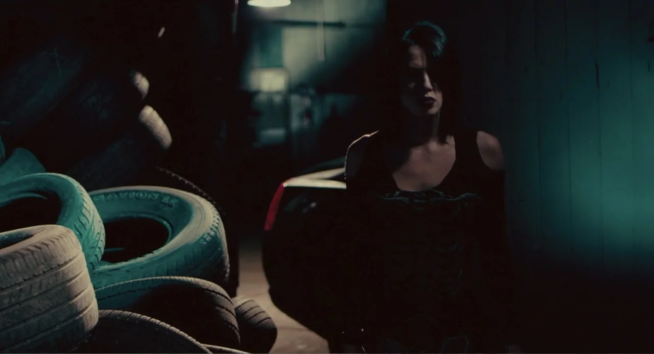 Terrifier (2018) movie still: medium — A dark, cluttered garage space features a large stack of discarded tires on the left. A young woman stands in…